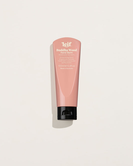 A pink 100ml hand balm tube with white lettering and a black flip-top cap is shown upright against a pale background with a soft shadow.