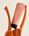 A pink hand balm tube with white lettering and a black cap is displayed at an angle between two glossy orange sculptural forms.