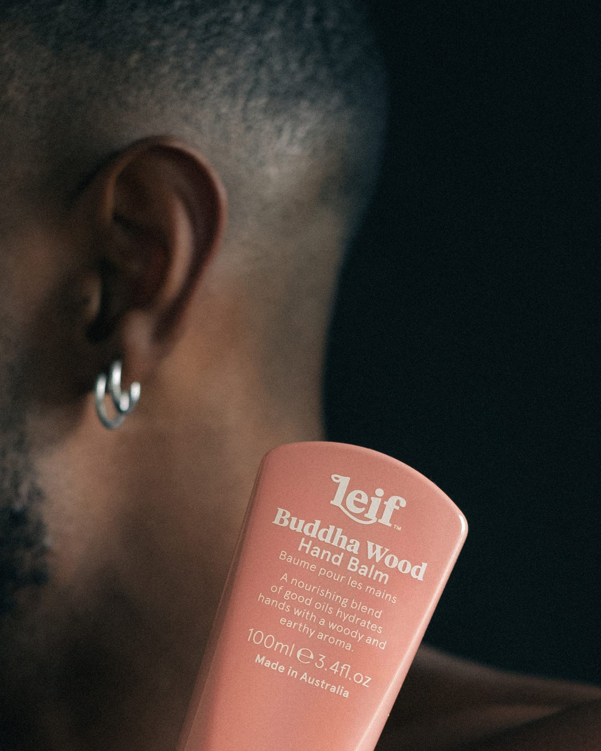 1. A close-up shows a pink hand balm tube with white lettering held beside a person's face and silver hoop earring against a dark background.
