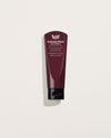 A burgundy 100ml hand balm tube with white lettering and a black flip-top cap stands upright against a pale background, casting a soft shadow.