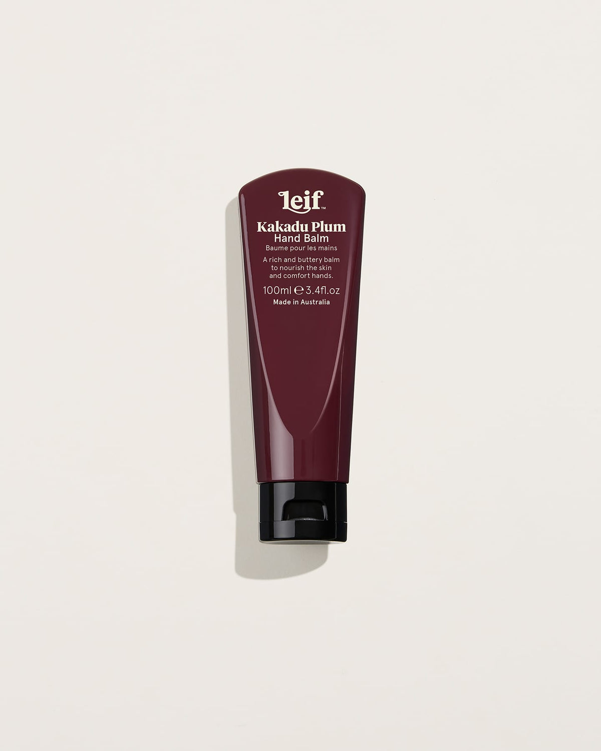 A burgundy 100ml hand balm tube with white lettering and a black flip-top cap stands upright against a pale background, casting a soft shadow.