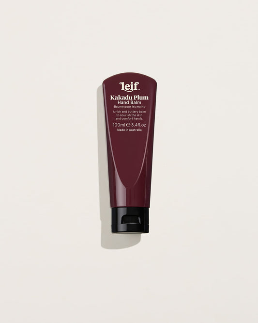 A burgundy 100ml hand balm tube with white lettering and a black flip-top cap stands upright against a pale background, casting a soft shadow.