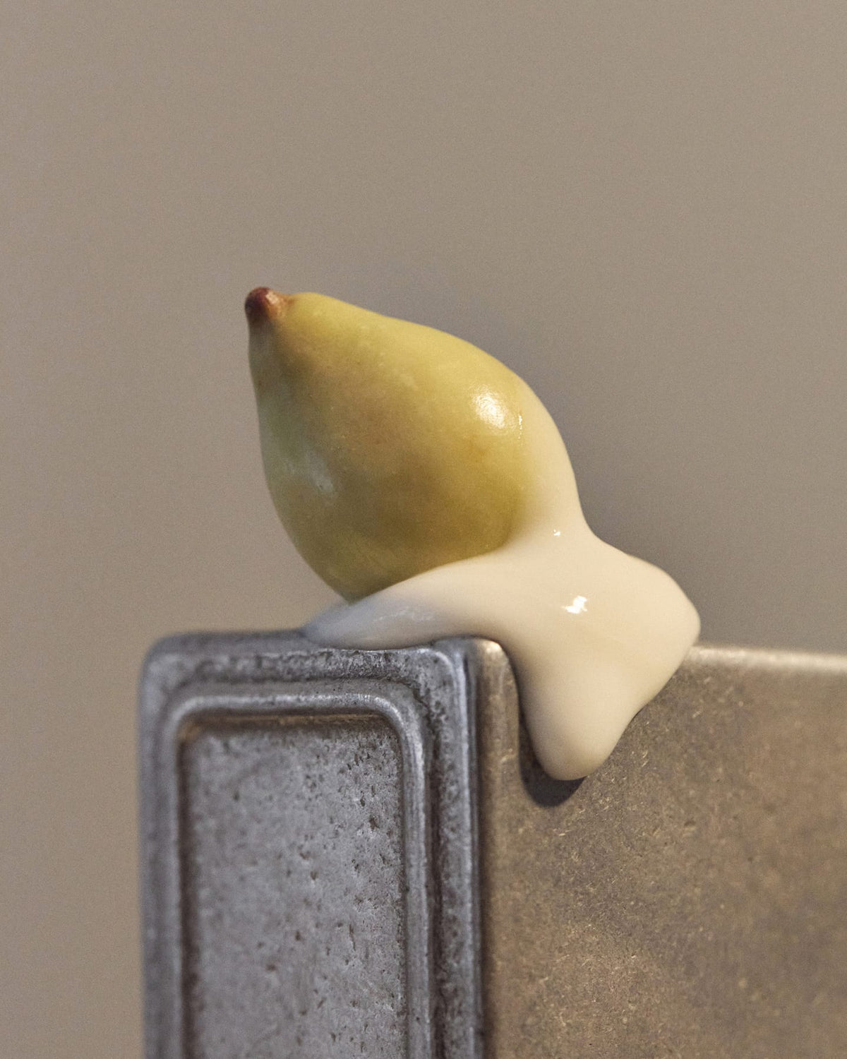 A close-up shows a pale creamy balm draped over the edge of a textured grey surface, with a small yellow-green fruit positioned above it.