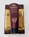 A burgundy hand balm tube with white lettering and a black cap rests vertically on a reflective gold-toned surface.