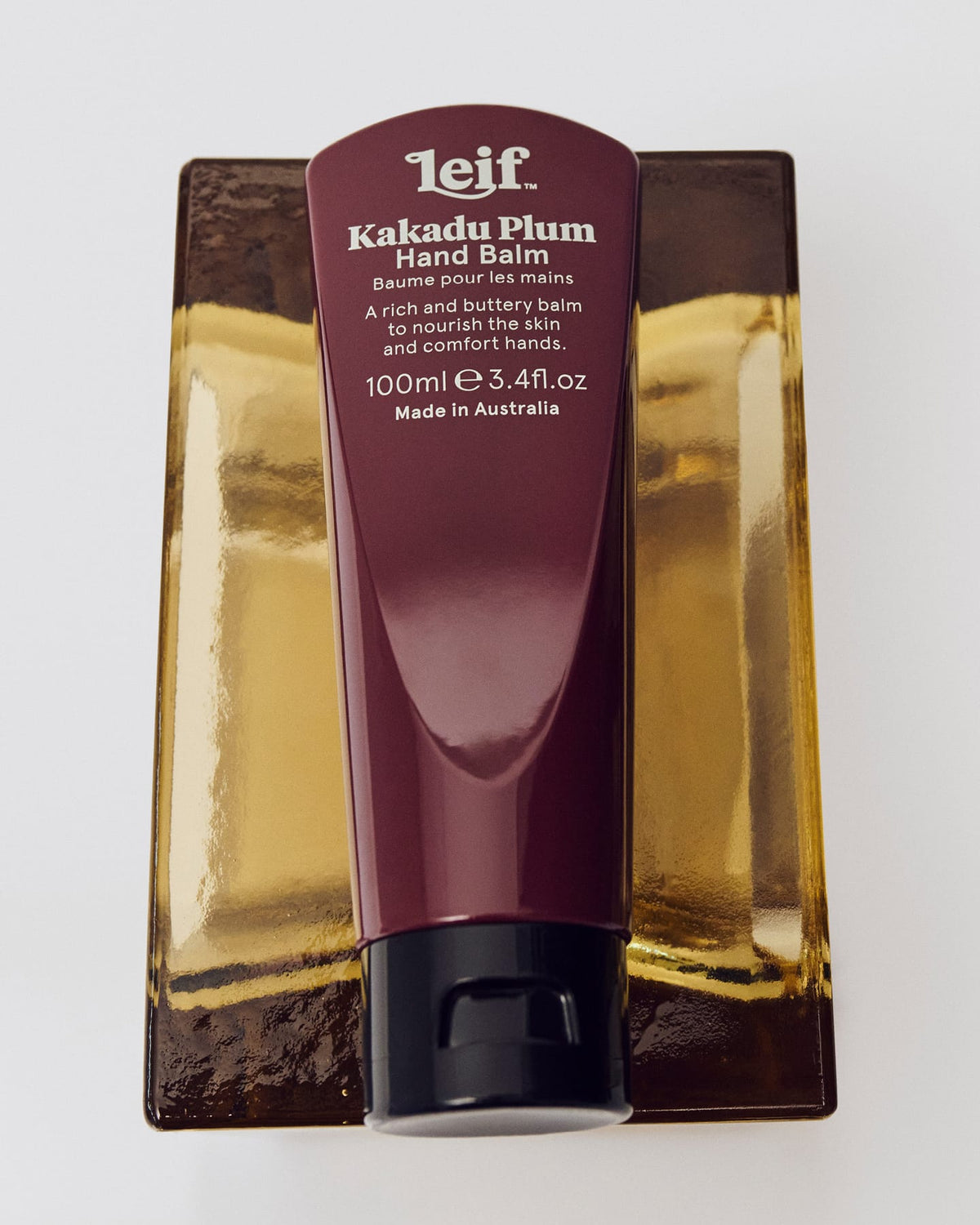 A burgundy hand balm tube with white lettering and a black cap rests vertically on a reflective gold-toned surface.