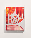 A limited-edition gift box featuring bold red, orange and cream abstract artwork with large white “HOLIDAY” lettering across the front.
