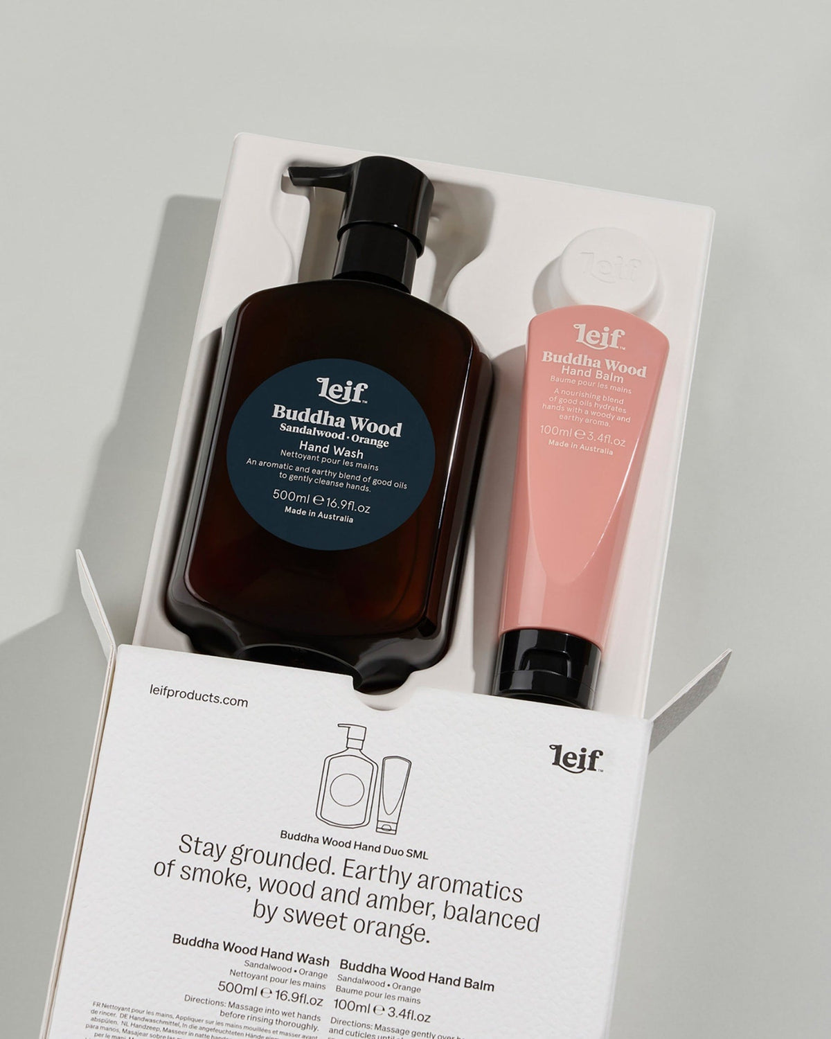 An open white gift box displays a dark hand wash bottle with a black pump and a pink hand balm tube, with printed product details on the packaging.