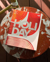 A colourful gift box featuring red, orange and cream abstract artwork rests on a round red-and-cream marble table outdoors.