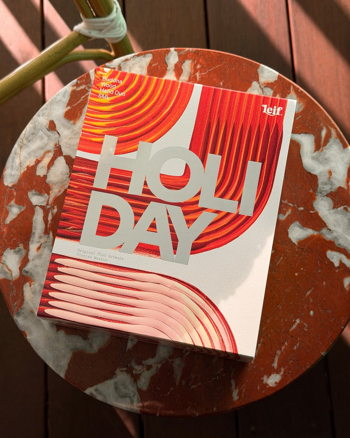 A colourful gift box featuring red, orange and cream abstract artwork rests on a round red-and-cream marble table outdoors.