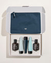 An open white gift box containing five body and hair care products in dark brown, pale turquoise and navy packaging, with a navy zip pouch in the lid.