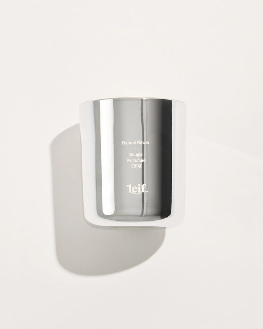 A chrome glass candle vessel with pale wax is shown from the front against a light background, displaying the Flannel Flower label.