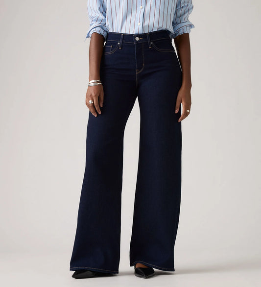 Model wearing the Darkest Sky wide-leg jeans with a blue striped shirt, showing the high-rise waist, five-pocket styling, dark indigo wash and full wide-leg silhouette in a cropped front view.