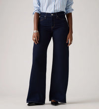 Model wearing the Darkest Sky wide-leg jeans with a blue striped shirt, showing the high-rise waist, five-pocket styling, dark indigo wash and full wide-leg silhouette in a cropped front view.