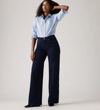 Model wearing the dark indigo wide-leg jeans with a blue striped shirt and black pointed shoes, showing the high-rise waist, fitted hip and full-length wide legs in a full-length front view.