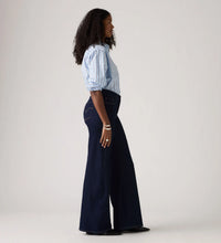 Model wearing the Darkest Sky wide-leg jeans with a blue striped shirt and black pointed shoes, showing the high-rise waist, fitted hip, back pocket detailing and full-length wide-leg silhouette in a side view.