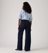 Model wearing the dark indigo wide-leg jeans with a blue striped shirt, showing the back pockets, curved back yoke, fitted waist and full-length wide-leg silhouette in a back view.