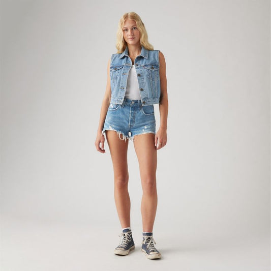Model wearing light blue high-rise denim shorts with a button closure, five-pocket design, distressed details and frayed hem, styled with a white tank top, sleeveless denim vest and high-top sneakers in a full-length front view.
