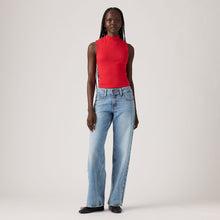 A model stands against a light gray studio background wearing a bright red sleeveless mock-neck top and light-wash blue jeans.