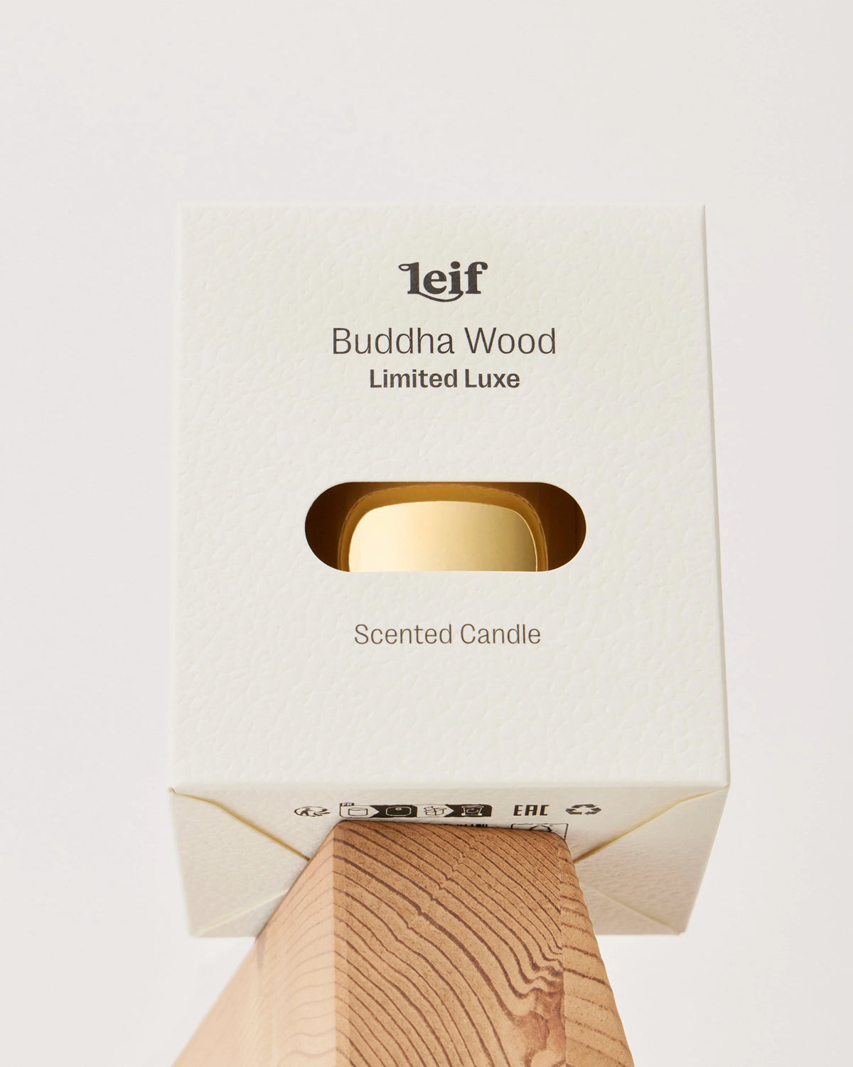 A cream Buddha Wood Limited Luxe candle box with a window revealing the gold vessel is displayed on a wooden block.