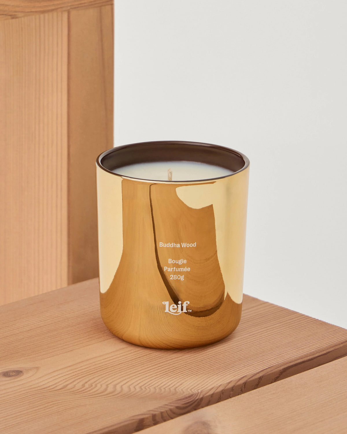 A gold glass Buddha Wood candle with pale wax and a visible wick sits on a wooden shelf beside a timber wall.