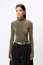 Woman wearing a green turtleneck sweater and black pants with a patterned belt on a white background