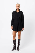 A model wears a black long-sleeve mini dress with a pointed collar and knee-high boots in a front studio view.