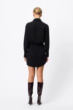 A model wears the black mini dress in a rear studio view showing the back yoke and curved hem.
