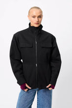 A model wearing a black zip-up jacket with a stand-up collar, two flap chest pockets, and ribbed cuffs, paired with blue jeans against a light gray studio background.
