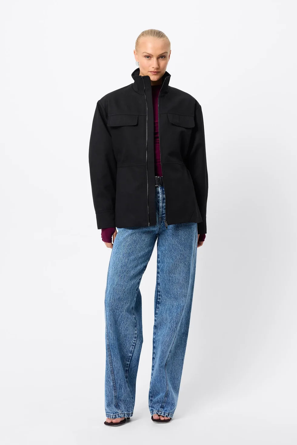 A model wears the black zip-front anorak with blue jeans in a full-length front studio view.

