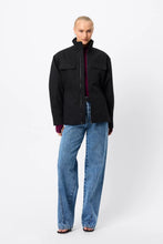 A model wears the black zip-front anorak with blue jeans in a full-length front studio view.
