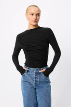 A model wearing a black long-sleeve top with ruching and a boat neckline, paired with blue jeans, against a light gray background.