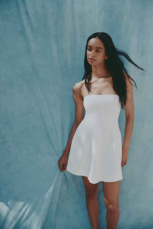 White mini dress shown on a model standing outdoors against a blue backdrop, with a strapless neckline and short flared skirt clearly visible.