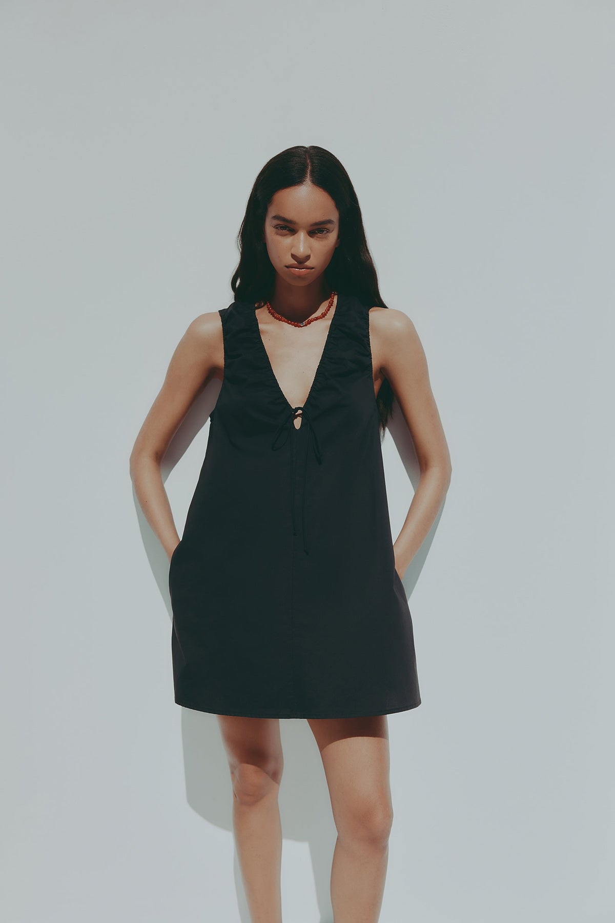 A model wears a black mini dress with a deep V neckline, gathered shoulders and a front tie while standing outdoors against a pale blue background.