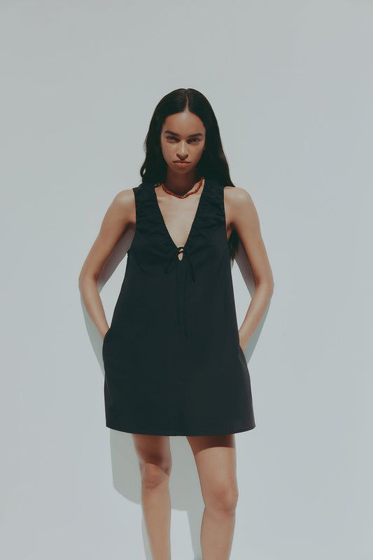 A model wears a black mini dress with a deep V neckline, gathered shoulders and a front tie while standing outdoors against a pale blue background.