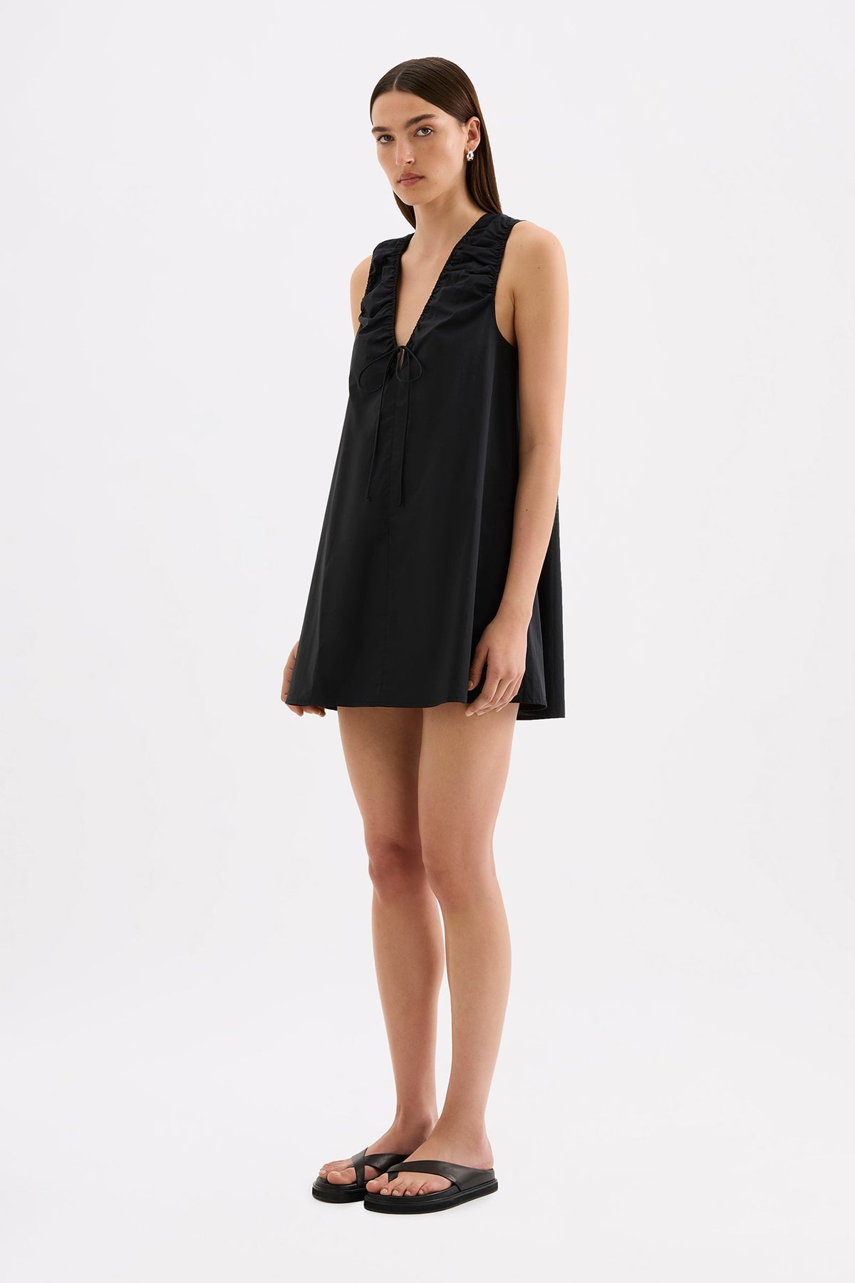 The model wears the black mini dress in a three-quarter front view, showing the deep V neckline, front tie and relaxed A-line shape.