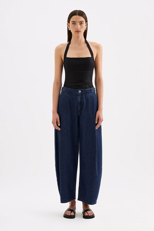 A model wears indigo blue wide-leg denim pants with deep pleats and a high waist in a full front studio view.