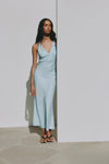 A model wears a Bermuda blue maxi dress with a V neckline and thin straps while standing against a pale wall outdoors.