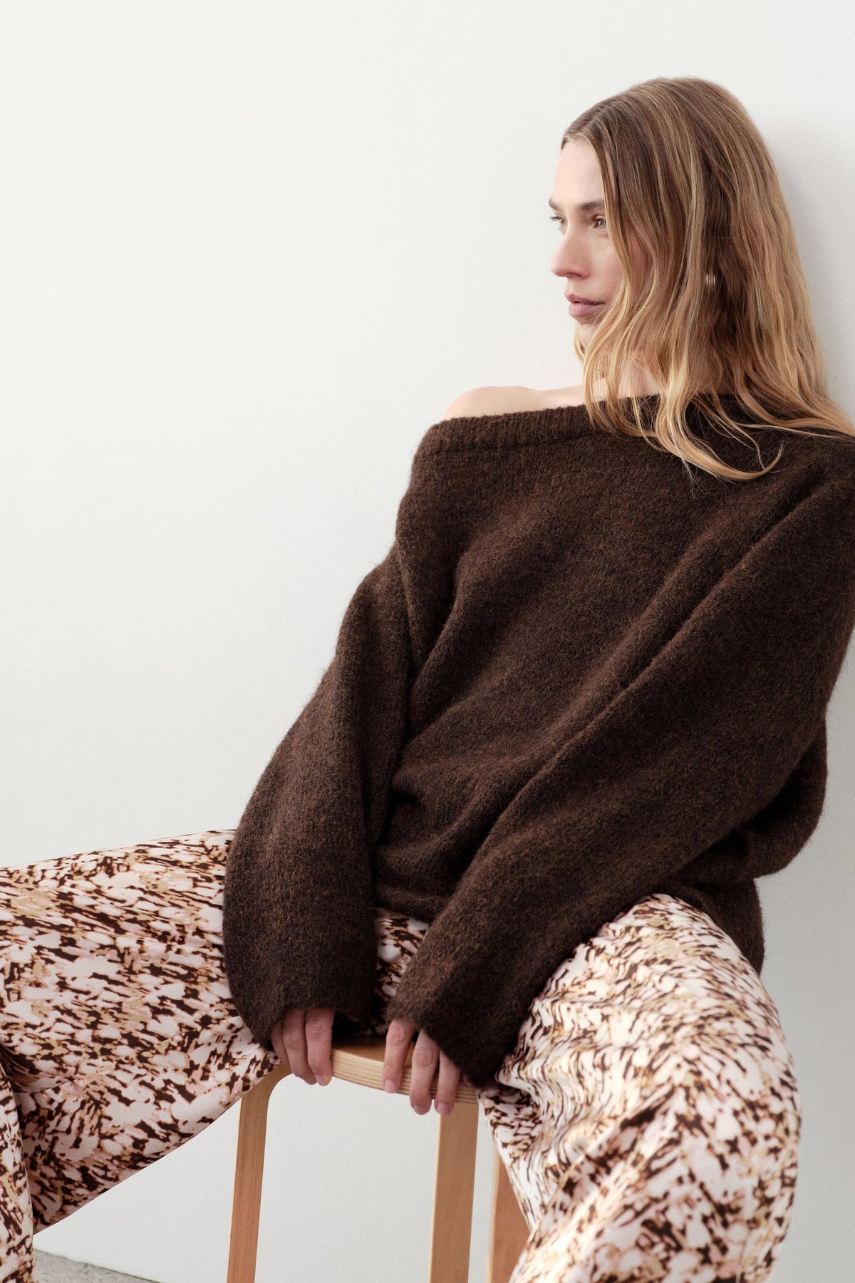 Woman seated on a stool wearing an oversized chocolate off-shoulder knit with patterned trousers.