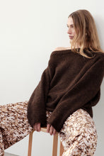 Woman seated on a stool wearing an oversized chocolate off-shoulder knit with patterned trousers.