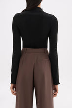 Back view of the black long sleeve top styled with brown tailored trousers against a white studio background.