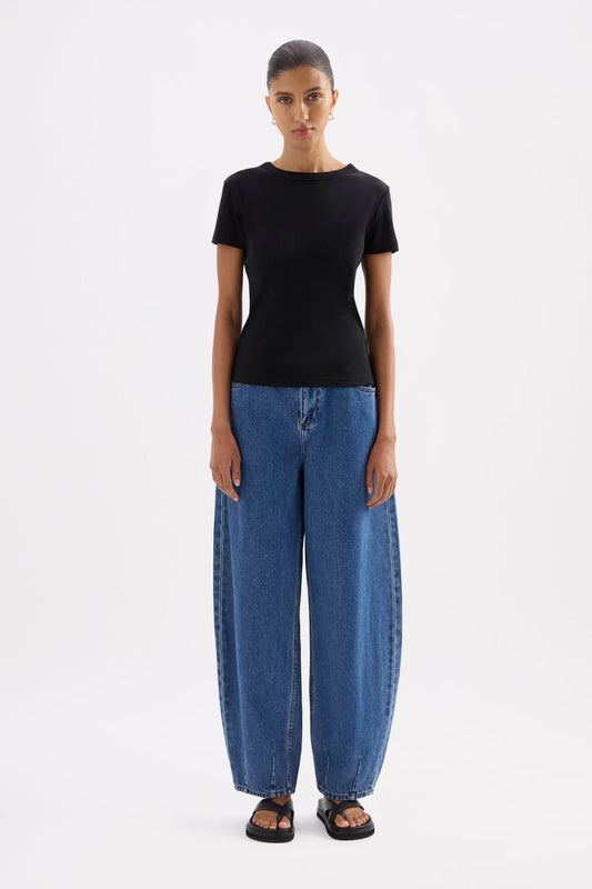 Full-length front view of the fitted black tee styled with relaxed blue jeans and black sandals in a white studio.