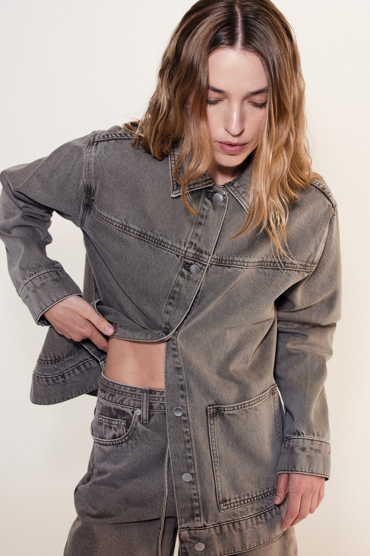 Woman seated wearing the stone grey denim jacket partially buttoned with matching jeans against a neutral studio background.