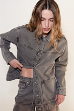 Woman seated wearing the stone grey denim jacket partially buttoned with matching jeans against a neutral studio background.