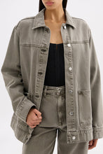 Model wearing the stone grey denim jacket open over a black top with matching jeans in a close front pose.