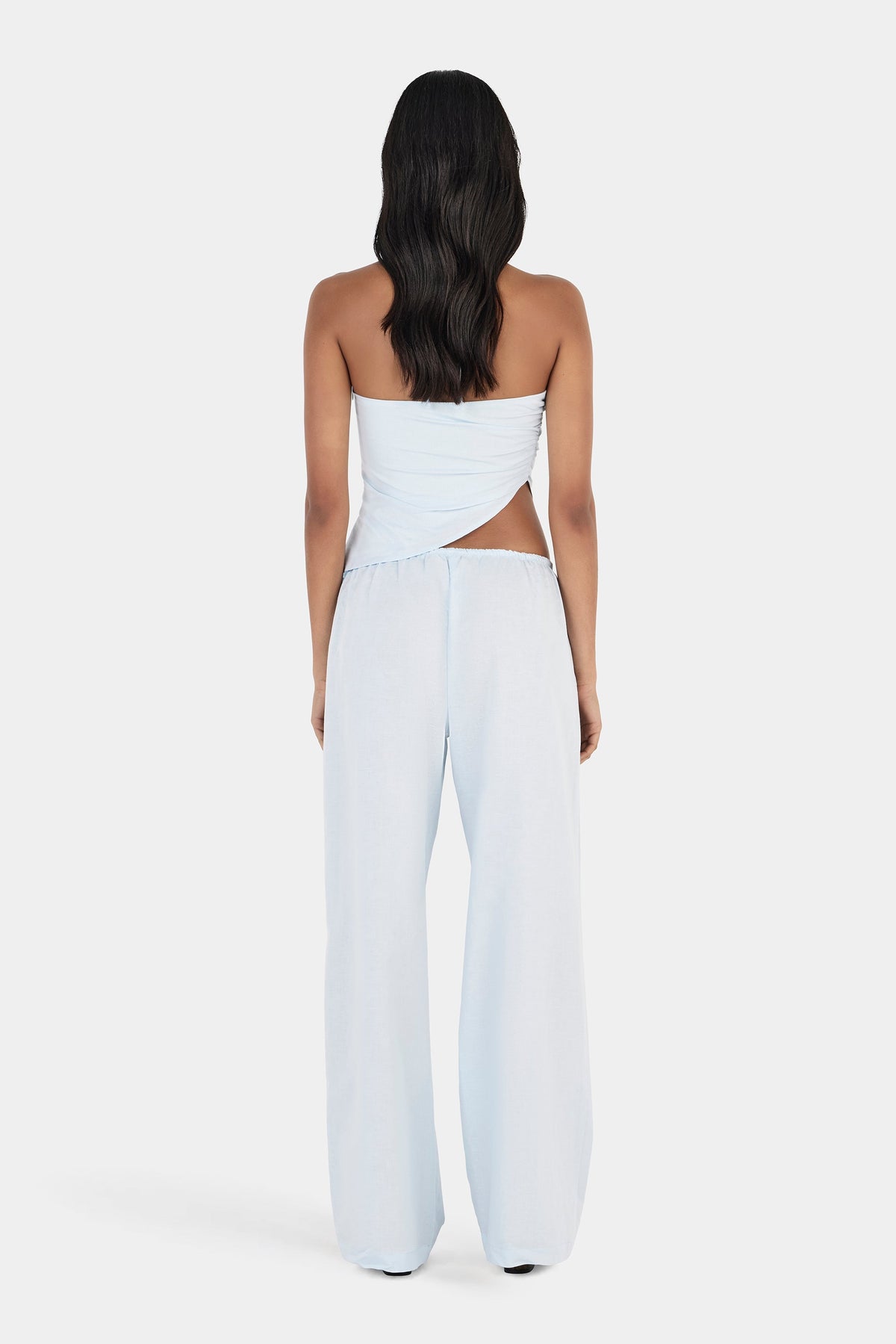 A rear view of a model wearing a pale blue top with a wrapped asymmetrical back, slim halterneck straps and an angled hem, paired with matching wide-leg trousers.
