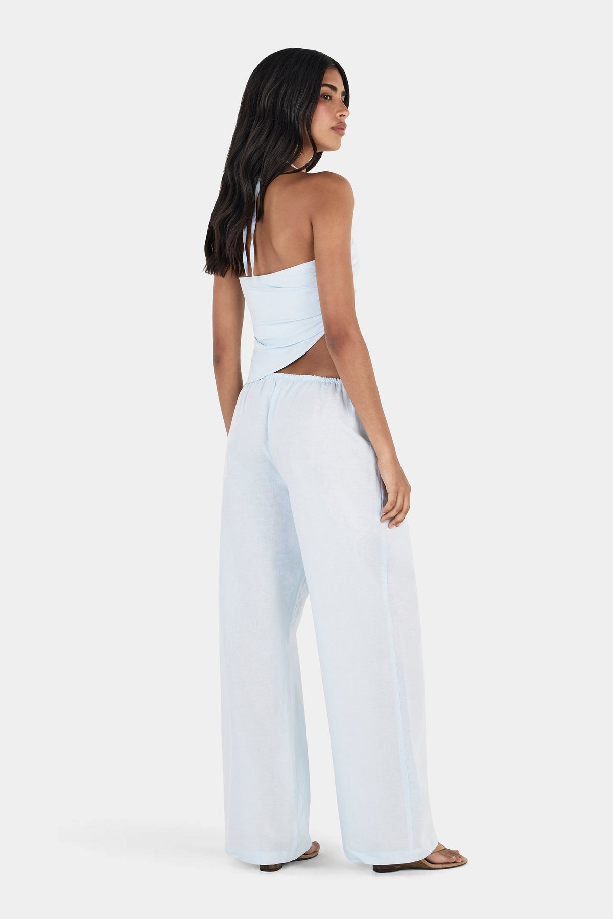 A model looking over her shoulder in a pale blue asymmetrical top with a low open back and diagonal hem, styled with matching wide-leg trousers in a rear three-quarter view.