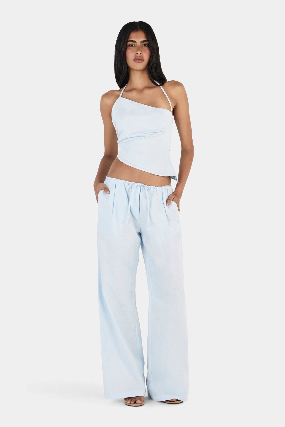 A model posing with her hands in the pockets of matching pale blue wide-leg trousers, wearing an asymmetrical top with thin halterneck straps and a diagonal hem.