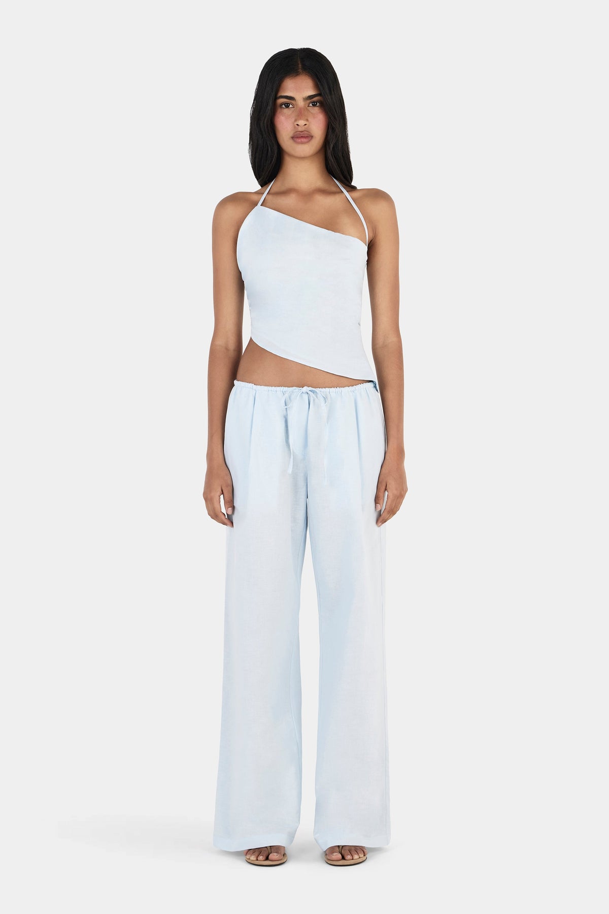 A front view of a model wearing a pale blue asymmetrical halterneck top with a slanted neckline and angled hem, paired with matching full-length wide-leg trousers.