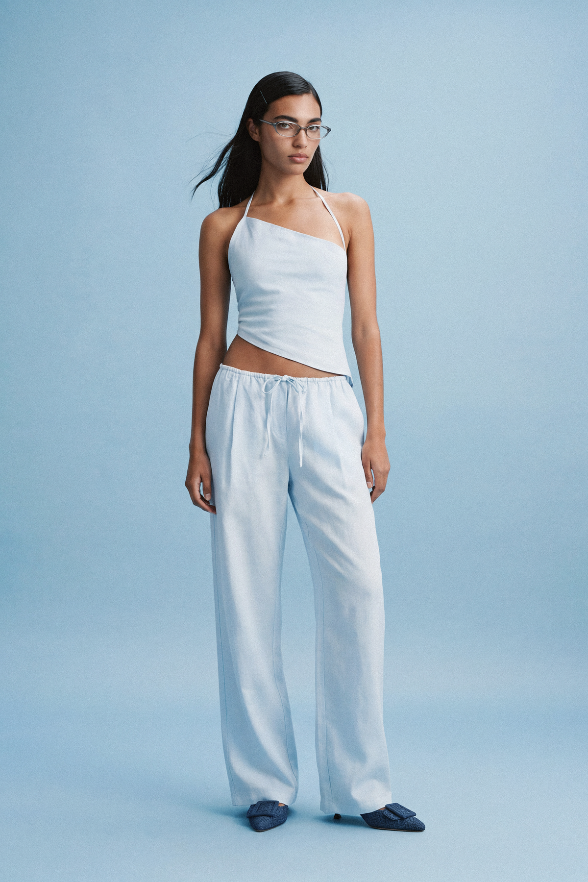 A model wearing a pale blue asymmetrical top and matching drawstring trousers against a blue studio background, styled with blue flats and clear-framed glasses.