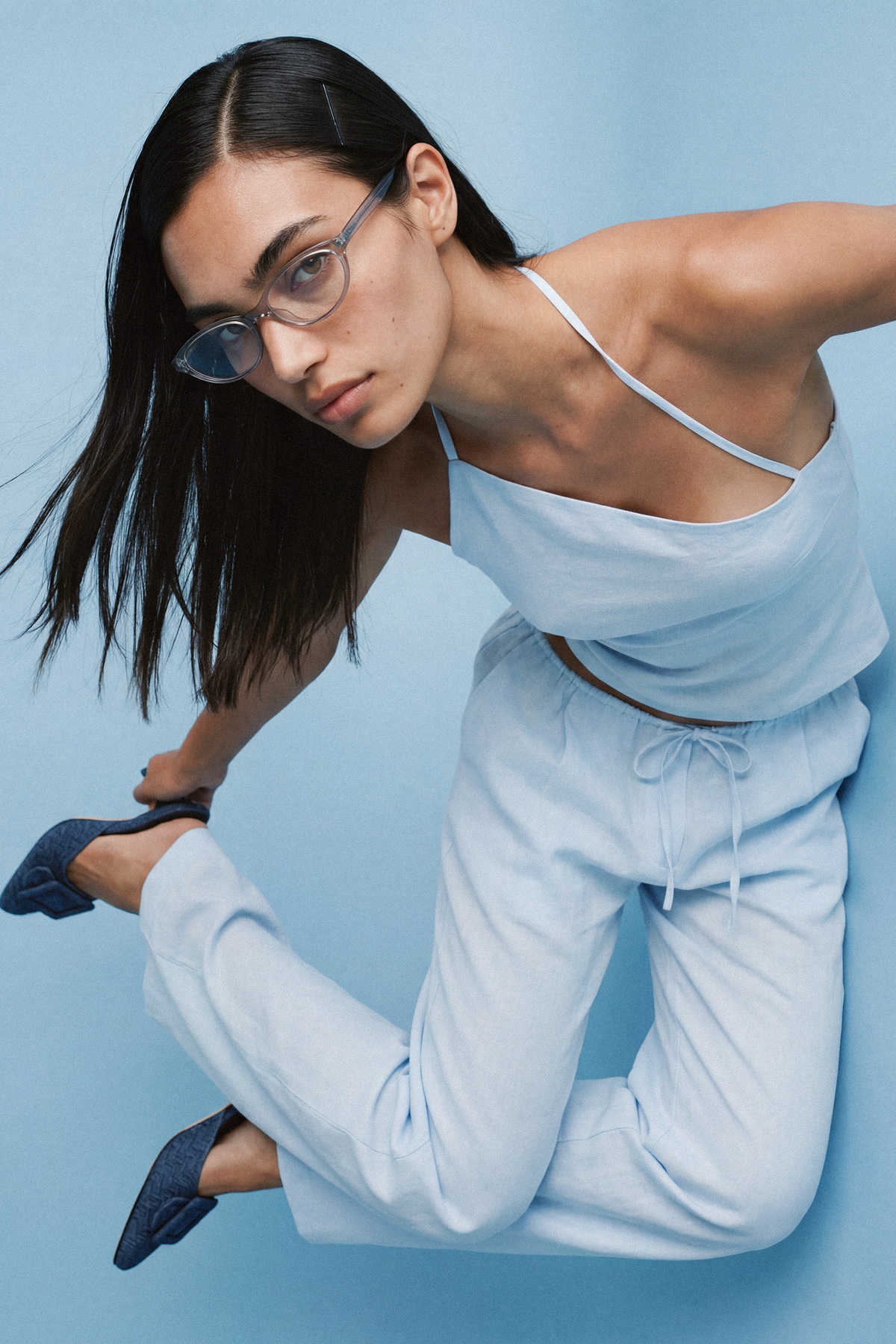  A close-up of a model leaning forward in a pale blue asymmetrical top with thin halterneck straps, showing the diagonal neckline and fitted bodice, paired with matching drawstring trousers.